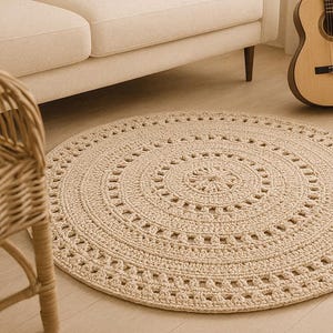 Op de afbeelding: Een ronde, beige gehaakte vloerkleed met een gedetailleerd cirkelvormig patroon. Het kleed ligt op een lichte houten vloer, in de buurt van een beige bank, een rieten stoel en een gitaar. De diameter van het kleed is ongeveer 152 cm.