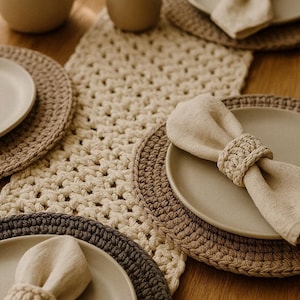 May include: A dining table arrangement with crochet placemats, plates, and napkins in neutral tones. The placemats are in beige, brown, and grey. Napkins are held by matching napkin rings. A teapot and cup are in the background.