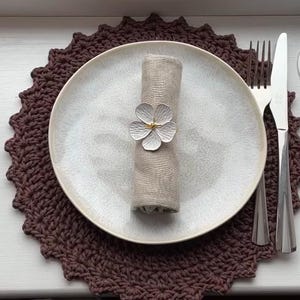 May include: A place setting with a brown crocheted placemat, a white plate, and a rolled beige napkin secured with a flower-shaped napkin ring. Silverware and a wine glass are also present.