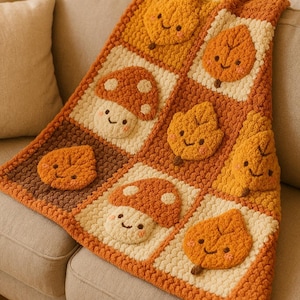 Könnte beinhalten: Handgefertigte Häkeldecke mit sich wiederholendem Muster aus lächelnden Pilzen und Blättern in Orange, Braun und Creme. Die Decke liegt über einem beigen Sofa und zeigt ihr strukturiertes Design und ihr herbstliches Thema.