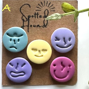 May include: Set of five colorful, round clay faces with different expressions: sad, neutral, winking, angry, and smiling. The faces are in shades of blue, yellow, purple, and pink. The text "Spotted Hound" and "Set A" are visible on the background.