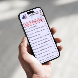 May include: A smartphone screen shows a semi-annual cleaning schedule. The list includes tasks such as washing walls, cleaning appliances, and decluttering. The phone is held in a hand, with the text in black against a white background.