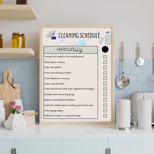 May include: A framed cleaning schedule titled "CLEANING SCHEDULE" with the word "ANNUALLY" in large text. The schedule lists tasks like washing windows and cleaning gutters. The background shows a kitchen setting with various utensils.