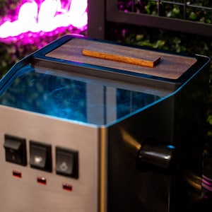 Puede incluir: Un electrodoméstico de acero inoxidable y negro con una tapa de madera y una pequeña pieza de acento de madera. El electrodoméstico tiene tres interruptores negros y un asa. El fondo incluye un letrero de neón y vegetación.