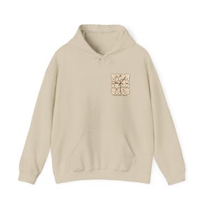 Puede incluir: Una sudadera con capucha beige con un bolsillo delantero y una capucha con cordón. La sudadera con capucha presenta el texto "TIME FEVER STOPS" en una fuente retro en la parte superior izquierda del pecho. El texto es de color marrón.