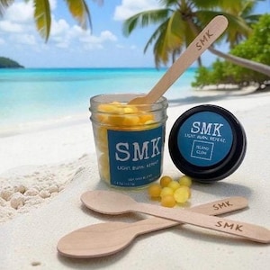 Puede incluir: Un tarro y un recipiente negro con el texto "SMK" y "LIGHT BURN, REPEAT" en una playa arenosa. El tarro contiene esferas amarillas y está acompañado de cucharas de madera. El fondo presenta una escena de playa tropical con palmeras y agua azul.