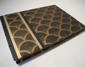 May include: A handmade journal with a black cover and a gold art deco fan pattern. The spine is black with visible stitching. A gold stripe runs down the center of the journal. The journal is open, revealing the inside pages.