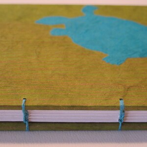 Pu&ograve; includere: Un diario verde con una silhouette di tartaruga blu sulla copertina. Il diario ha una spina dorsale bianca e chiusure a nastro blu.