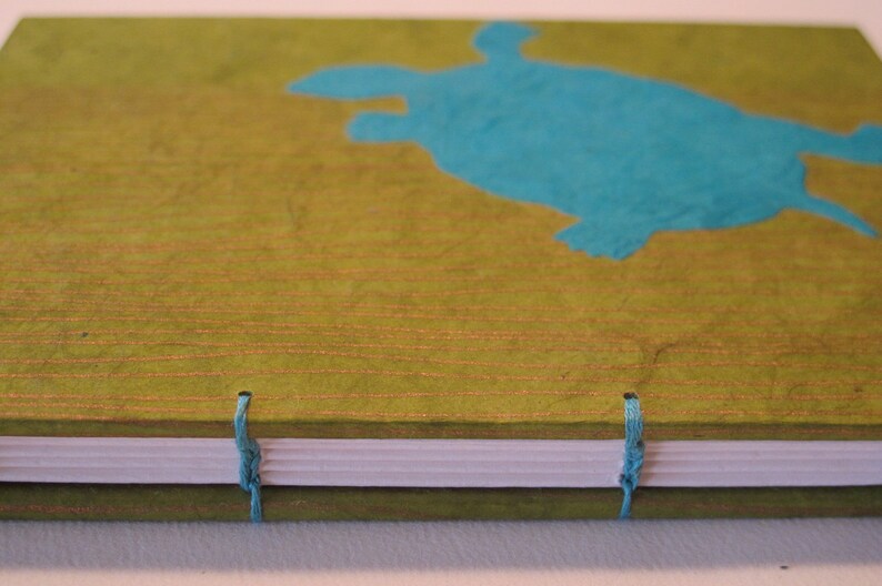 Pu&ograve; includere: Un diario verde con una silhouette di tartaruga blu sulla copertina. Il diario ha una spina dorsale bianca e chiusure a nastro blu.