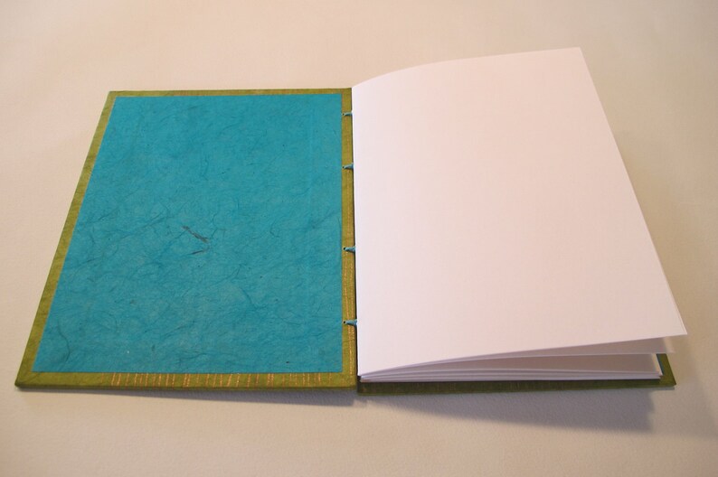 Pu&ograve; includere: Un diario artigianale blu turchese con un bordo verde e pagine bianche. Il diario &egrave; aperto per mostrare le pagine vuote.