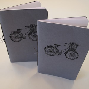 May include: Two gray notebooks with a bicycle illustration on the cover. The bicycle has a basket filled with flowers. The notebooks are open, revealing white pages.