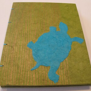 Pu&ograve; includere: Un diario verde con una silhouette di tartaruga di carta blu sulla copertina. Il diario ha una copertina testurizzata con linee dorate.
