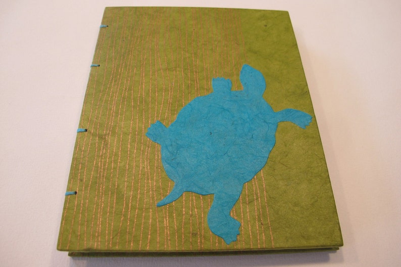 Pu&ograve; includere: Un diario verde con una silhouette di tartaruga di carta blu sulla copertina. Il diario ha una copertina testurizzata con linee dorate.