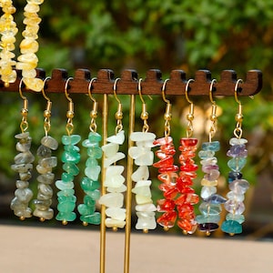 May include: A display of gemstone earrings in various colors, including green, red, and yellow. The earrings are suspended from a wooden rack with gold-colored hooks. The gemstones are irregularly shaped and strung together.