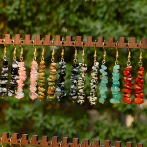May include: A display of gemstone earrings in various colors, including black, pink, green, and red. Each earring features a gold-colored hook and a strand of irregularly shaped stones. The earrings are displayed on a wooden rack against a blurred green background.