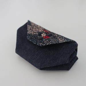 May include: A small, folded denim pouch with a patterned flap. The flap features a floral design with red accents and a black button closure. The pouch is a dark blue colour.