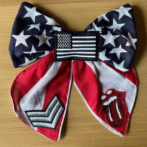 Op de afbeelding: Een stoffen haarstrik met een donkerblauwe achtergrond en witte sterren op de bovenste lussen. Het midden is versierd met een rechthoekige Amerikaanse vlag-patch. De onderste uiteinden tonen rode en witte strepen, een zilveren chevron-patch en een rode lippen-patch.