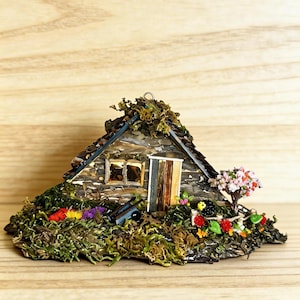 Puede incluir: Cabaña rústica en miniatura con techo y base cubiertos de musgo, con una pequeña puerta y ventanas. La cabaña está rodeada de coloridas flores en miniatura y un pequeño árbol con flores rosas. La cabaña está sobre una superficie de madera.