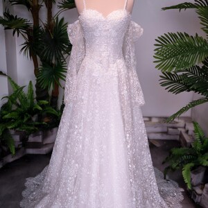 May include: A floor-length white wedding dress featuring intricate lace embroidery, a sweetheart neckline, thin spaghetti straps, and detachable off-the-shoulder long sleeves. The gown has a full skirt with a scalloped lace hem and a gold crown.