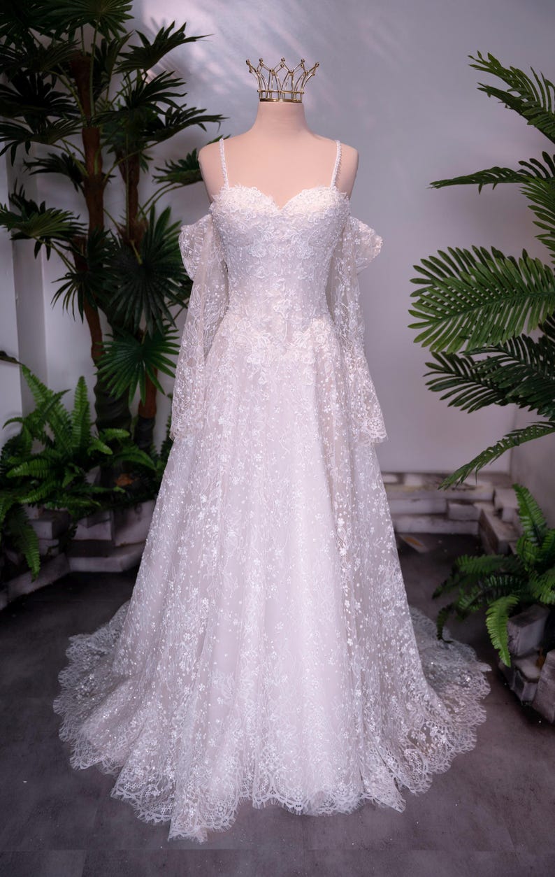 May include: A floor-length white wedding dress featuring intricate lace embroidery, a sweetheart neckline, thin spaghetti straps, and detachable off-the-shoulder long sleeves. The gown has a full skirt with a scalloped lace hem and a gold crown.