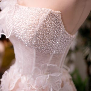 May include: A close-up of a bridal gown on a mannequin. The bodice is heavily embellished with small, white pearls. The dress features a sweetheart neckline and delicate, layered fabric with floral accents. The overall color is a soft, pale pink.