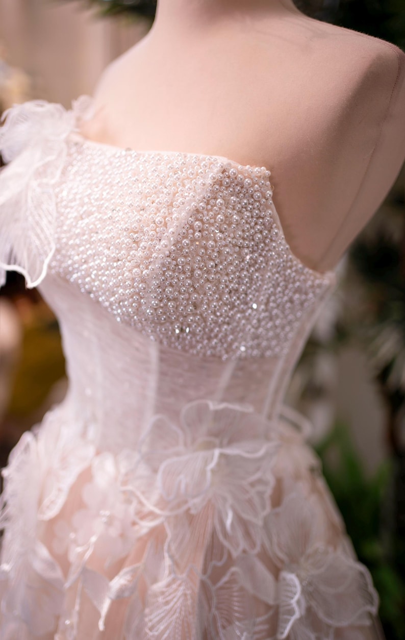 May include: A close-up of a bridal gown on a mannequin. The bodice is heavily embellished with small, white pearls. The dress features a sweetheart neckline and delicate, layered fabric with floral accents. The overall color is a soft, pale pink.