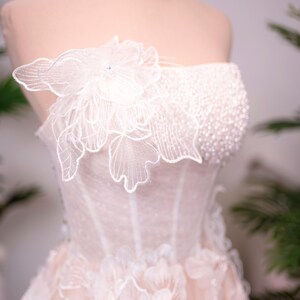 May include: A strapless, ivory-colored wedding dress on a mannequin. The bodice is embellished with small beads and a large, delicate floral applique. The skirt features layered floral appliques, creating a romantic and elegant look.