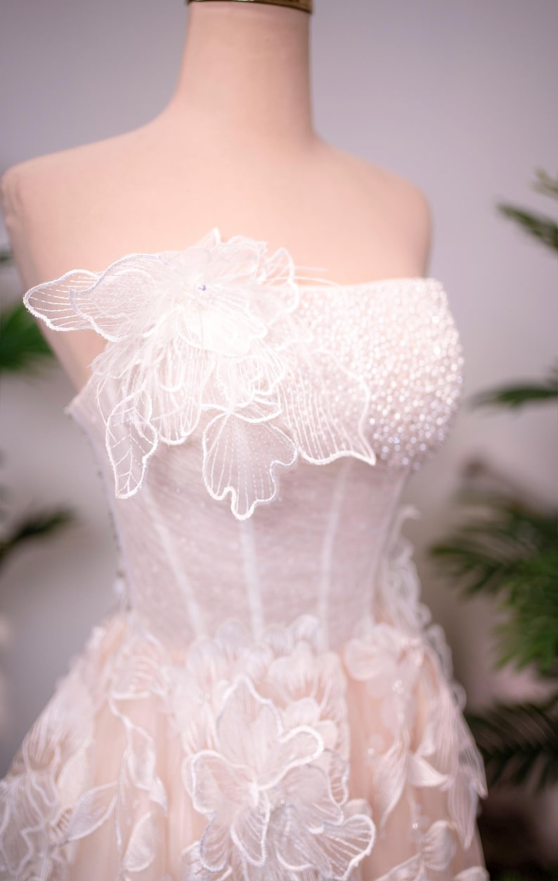 May include: A strapless, ivory-colored wedding dress on a mannequin. The bodice is embellished with small beads and a large, delicate floral applique. The skirt features layered floral appliques, creating a romantic and elegant look.