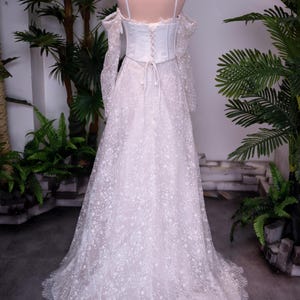 May include: A floor-length white gown featuring a corset-style bodice with lace-up back detailing, thin shoulder straps, and off-the-shoulder long sleeves. The skirt is made of sheer lace with a floral pattern and a scalloped hemline, topped with a crown.