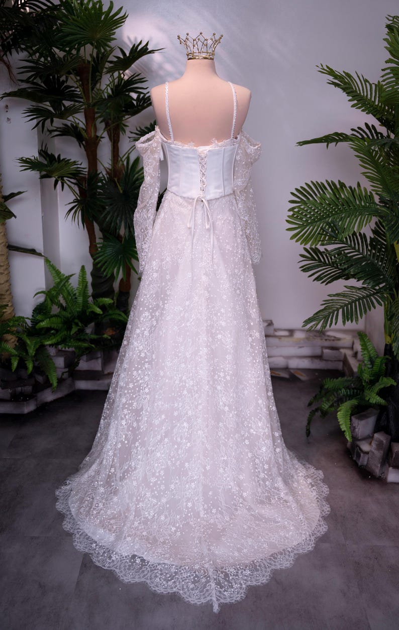 May include: A floor-length white gown featuring a corset-style bodice with lace-up back detailing, thin shoulder straps, and off-the-shoulder long sleeves. The skirt is made of sheer lace with a floral pattern and a scalloped hemline, topped with a crown.