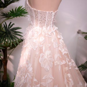 May include: A strapless wedding gown on a mannequin. The bodice is embellished with beads and sequins, transitioning into a flowing skirt adorned with large, embroidered floral appliques. The dress is a soft blush color.