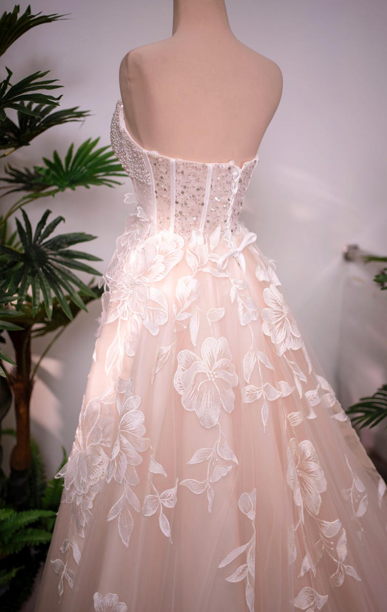 May include: A strapless wedding gown on a mannequin. The bodice is embellished with beads and sequins, transitioning into a flowing skirt adorned with large, embroidered floral appliques. The dress is a soft blush color.