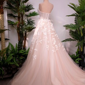 May include: A blush-colored wedding gown on a mannequin, featuring a strapless bodice with intricate beadwork and floral appliqu&eacute;s. The skirt is made of layers of soft tulle, and the train flows gracefully. A gold crown sits atop the mannequin's head.