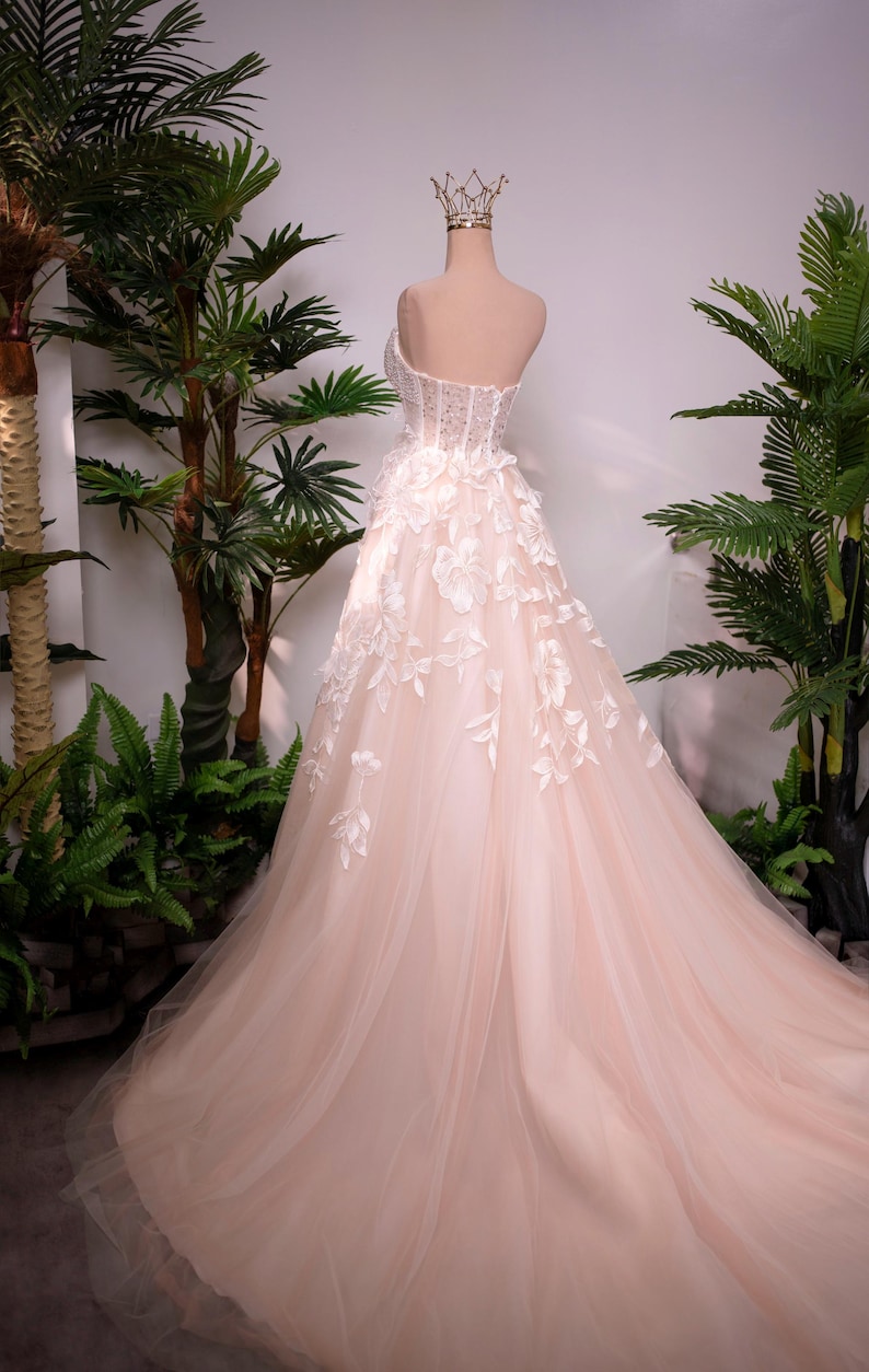 May include: A blush-colored wedding gown on a mannequin, featuring a strapless bodice with intricate beadwork and floral appliqu&eacute;s. The skirt is made of layers of soft tulle, and the train flows gracefully. A gold crown sits atop the mannequin's head.