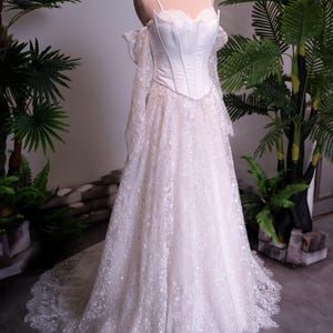 May include: A floor-length white gown featuring a structured corset bodice with thin straps and off-the-shoulder long lace sleeves. The skirt is made of intricate floral lace with a scalloped hemline, and the mannequin wears a gold crown.