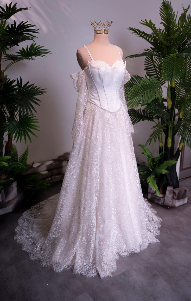 May include: A floor-length white gown featuring a structured corset bodice with thin straps and off-the-shoulder long lace sleeves. The skirt is made of intricate floral lace with a scalloped hemline, and the mannequin wears a gold crown.
