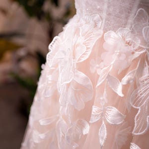 May include: Close-up of a wedding dress bodice. The dress features intricate white floral lace appliqu&eacute;s over a pale pink fabric. The lace detailing includes large flowers and delicate leaves, creating a romantic and elegant look.