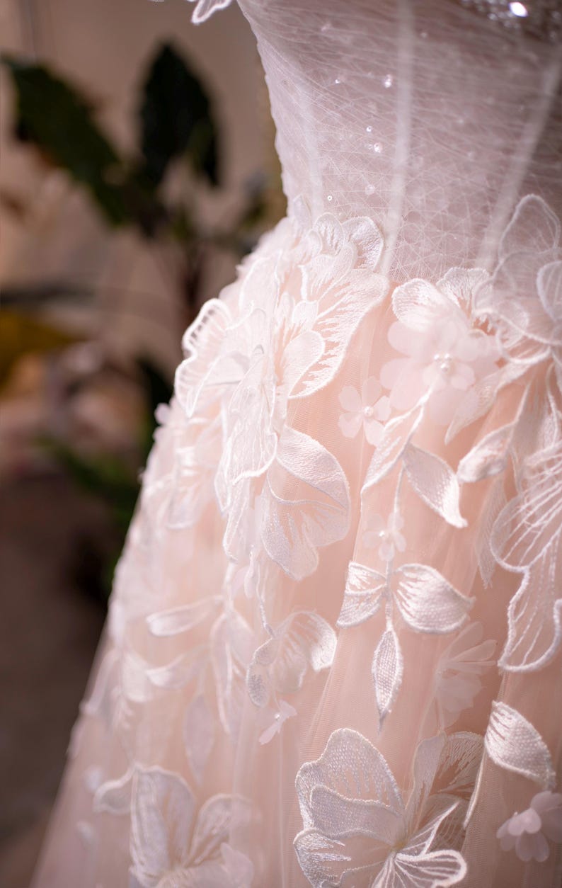May include: Close-up of a wedding dress bodice. The dress features intricate white floral lace appliqu&eacute;s over a pale pink fabric. The lace detailing includes large flowers and delicate leaves, creating a romantic and elegant look.