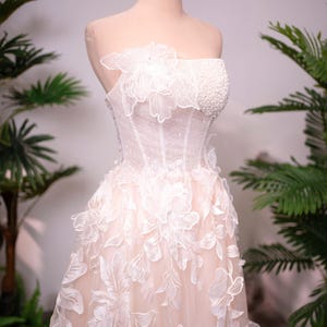 May include: A strapless, ivory wedding dress with floral appliques and a beaded bodice is displayed on a mannequin. A gold crown sits atop the mannequin's head. The dress features a layered skirt and delicate floral details.