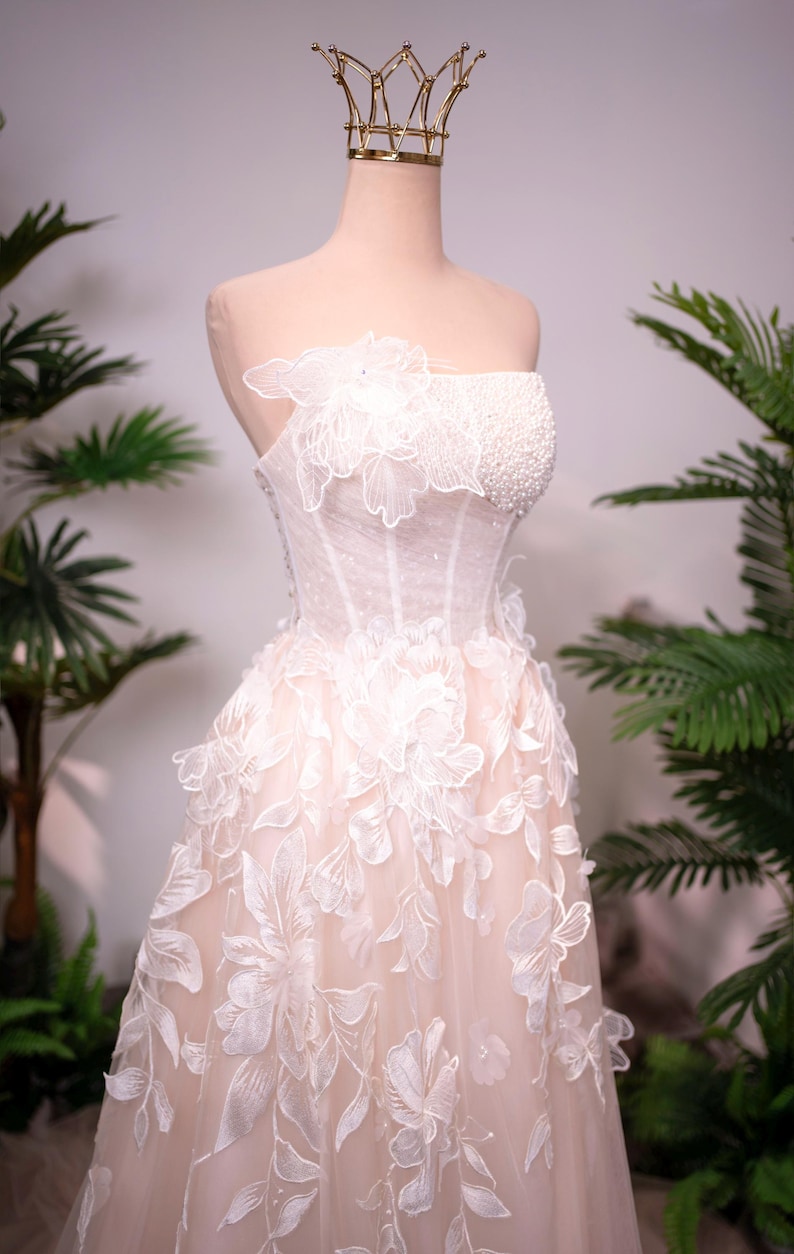 May include: A strapless, ivory wedding dress with floral appliques and a beaded bodice is displayed on a mannequin. A gold crown sits atop the mannequin's head. The dress features a layered skirt and delicate floral details.