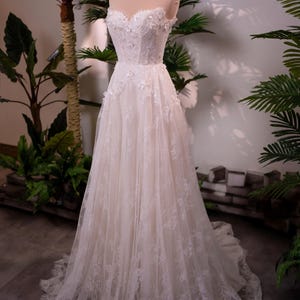 May include: A white wedding dress on a mannequin. The strapless gown features floral lace detailing on the bodice and skirt. A gold crown sits atop the mannequin's head. The dress is displayed in front of green plants.