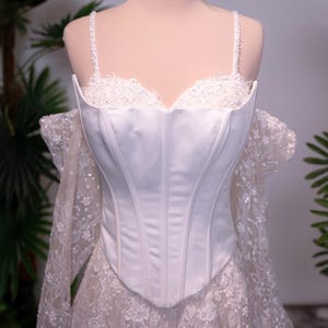 May include: A white corset bodice featuring a smooth satin finish and structured boning, paired with delicate lace off-the-shoulder sleeves and a matching lace skirt overlay. A gold metal crown sits atop the mannequin head.