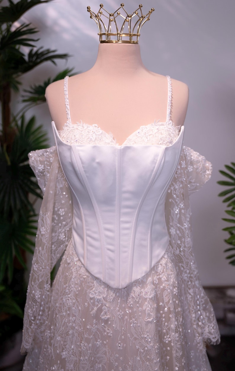 May include: A white corset bodice featuring a smooth satin finish and structured boning, paired with delicate lace off-the-shoulder sleeves and a matching lace skirt overlay. A gold metal crown sits atop the mannequin head.