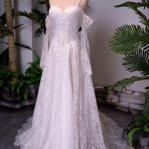 May include: A floor-length off-white lace gown featuring a sweetheart neckline, thin spaghetti straps, and detachable long lace sleeves. The dress is adorned with intricate floral embroidery and sequins, with a full skirt and a train.