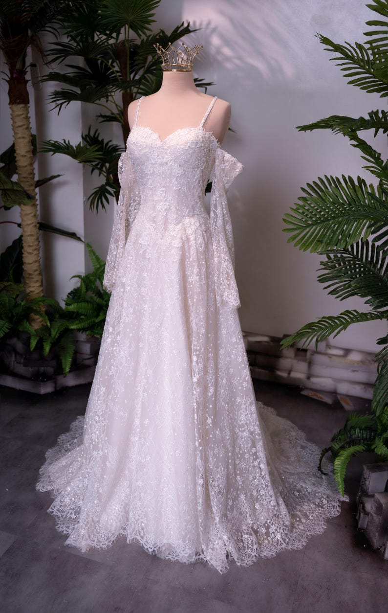 May include: A floor-length off-white lace gown featuring a sweetheart neckline, thin spaghetti straps, and detachable long lace sleeves. The dress is adorned with intricate floral embroidery and sequins, with a full skirt and a train.