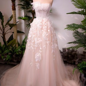 May include: A strapless blush-colored wedding dress on a mannequin. The bodice is embellished with delicate lace and beading, while the skirt features floral appliqu&eacute;s and a flowing tulle overlay. A gold crown sits atop the mannequin's head.