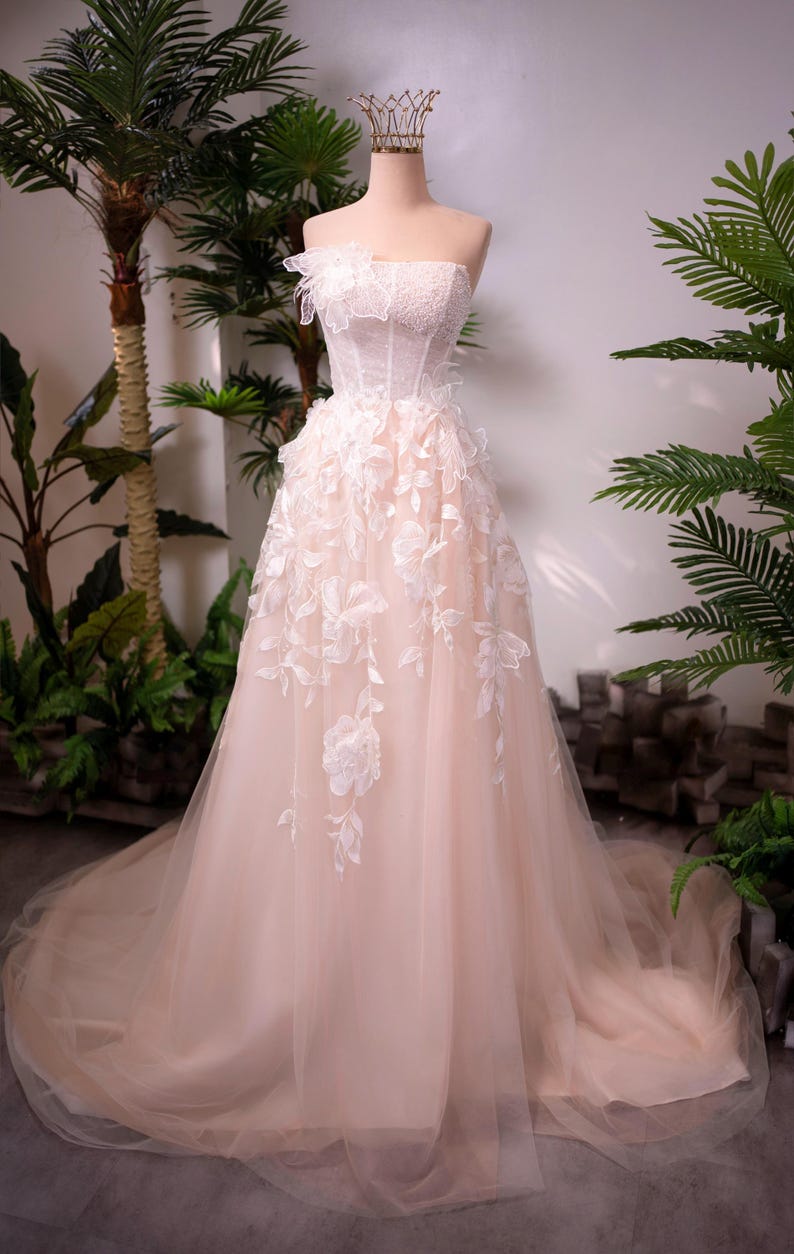 May include: A strapless blush-colored wedding dress on a mannequin. The bodice is embellished with delicate lace and beading, while the skirt features floral appliqu&eacute;s and a flowing tulle overlay. A gold crown sits atop the mannequin's head.