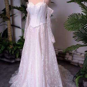 May include: A floor-length white gown featuring a structured corset bodice with thin straps and off-the-shoulder long lace sleeves. The skirt is made of intricate floral lace with a scalloped hemline, and a small gold crown sits atop the mannequin.