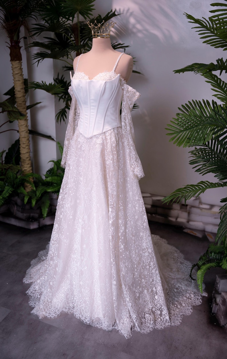 May include: A floor-length white gown featuring a structured corset bodice with thin straps and off-the-shoulder long lace sleeves. The skirt is made of intricate floral lace with a scalloped hemline, and a small gold crown sits atop the mannequin.