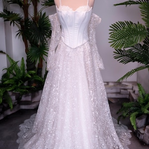 May include: A white floor-length gown featuring a structured corset bodice with thin straps and off-the-shoulder long lace sleeves. The skirt is made of layered white lace with a delicate floral pattern, and a gold crown sits atop the mannequin.